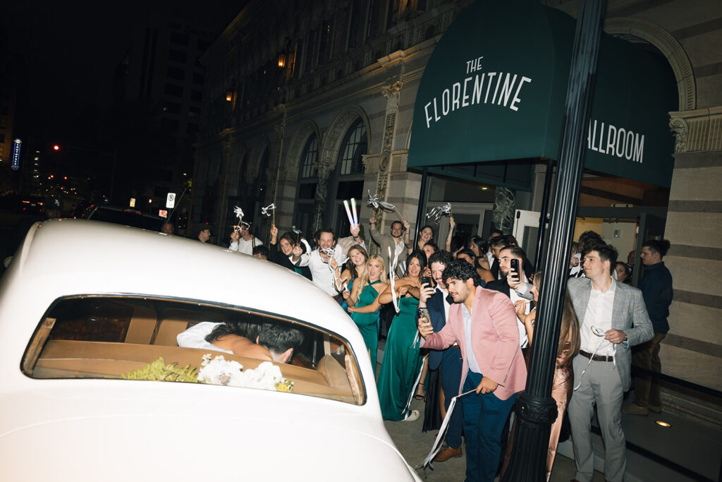 Wedding guests take photos of bride and groom as they drive away in their vintage getaway car